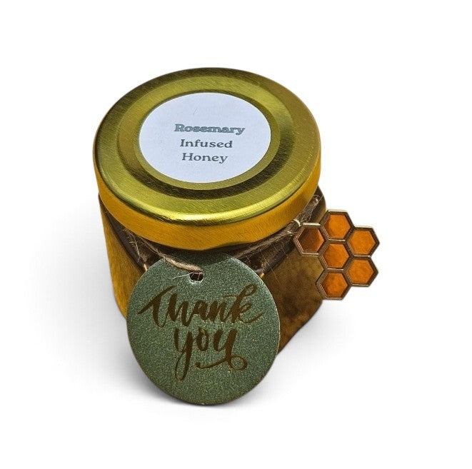 A small jar of Rosemary Infused Honey with a gold lid, a white label, and a green glittery 'thank you' tag wrapped around the jar, next to a honeycomb-shaped embellishment, on a white background.
