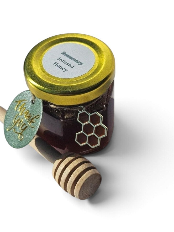 A jar of infused honey with a gold lid and a hanging green tag reading ' Rosemary Infused Honey,' shown with a wooden honey dipper on a white background.