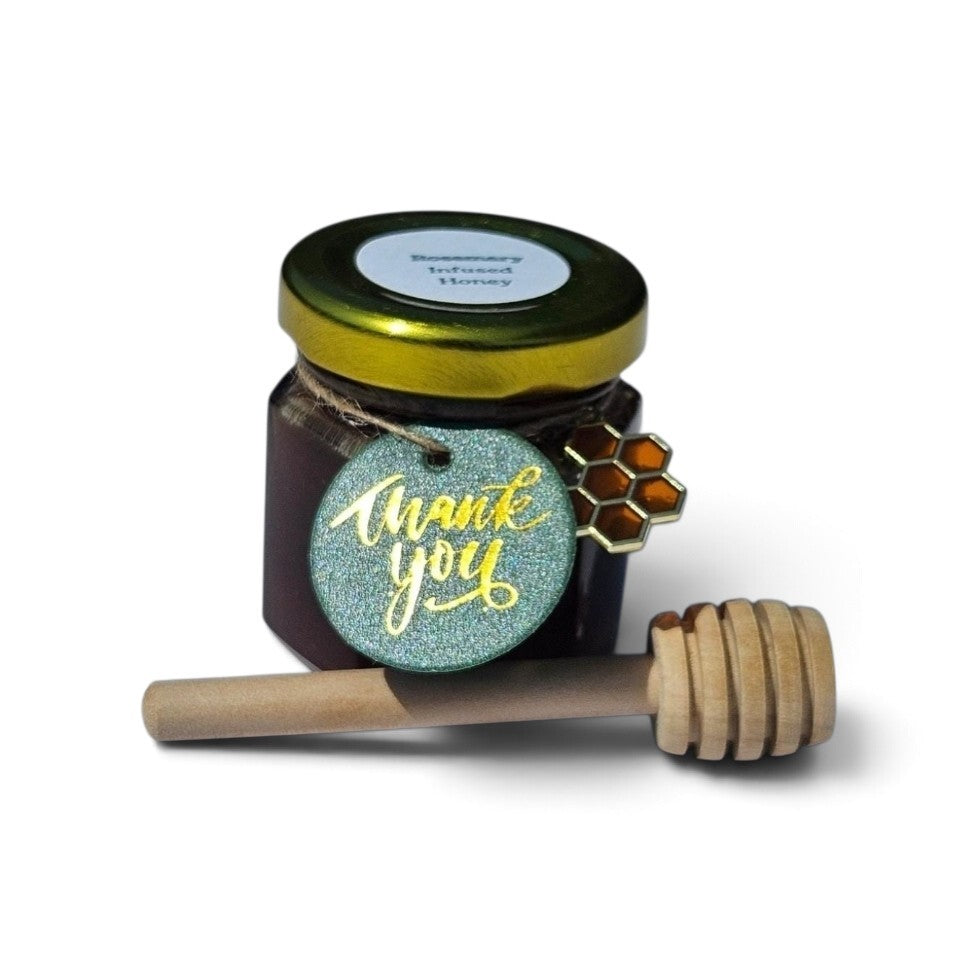 A sample jar of  Rosemary Infused honey with a gold lid, a wooden honey dipper, and a glittery green 'thank you' tag on a white background.
