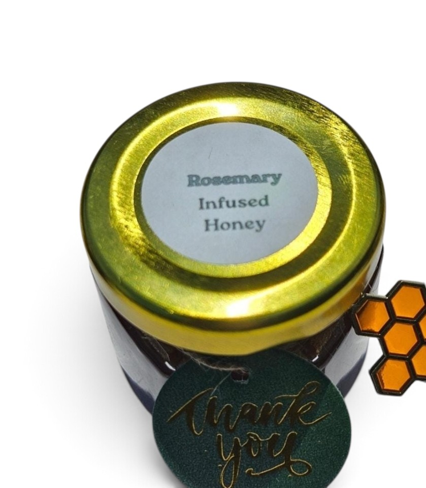 A small jar of Rosemary Infused Honey with a shiny gold lid and a round white label. A dark green circular 'thank you' tag with gold script is attached, and a small orange honeycomb-shaped tag is visible on the right, all on a white background.