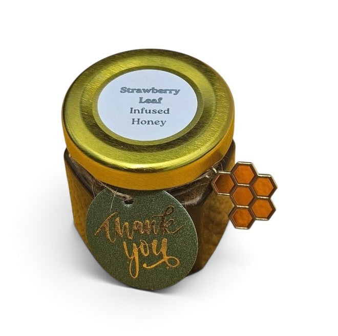 A sample jar of strawberry leaf infused honey with a gold lid, a white label, and a green 'thank you' tag with a gold honeycomb charm, shown on a white background.