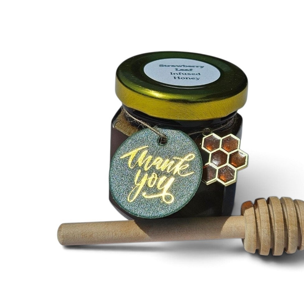 A sample jar of Strawberry Lemon Infused Honey with a gold lid, decorated with a glittery blue 'thank you' tag and a honeycomb charm, shown with a wooden honey dipper on a white background.