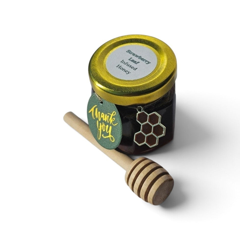 A small glass jar of strawberry leaf infused honey with a gold lid, shown with a wooden honey dipper on a white background.