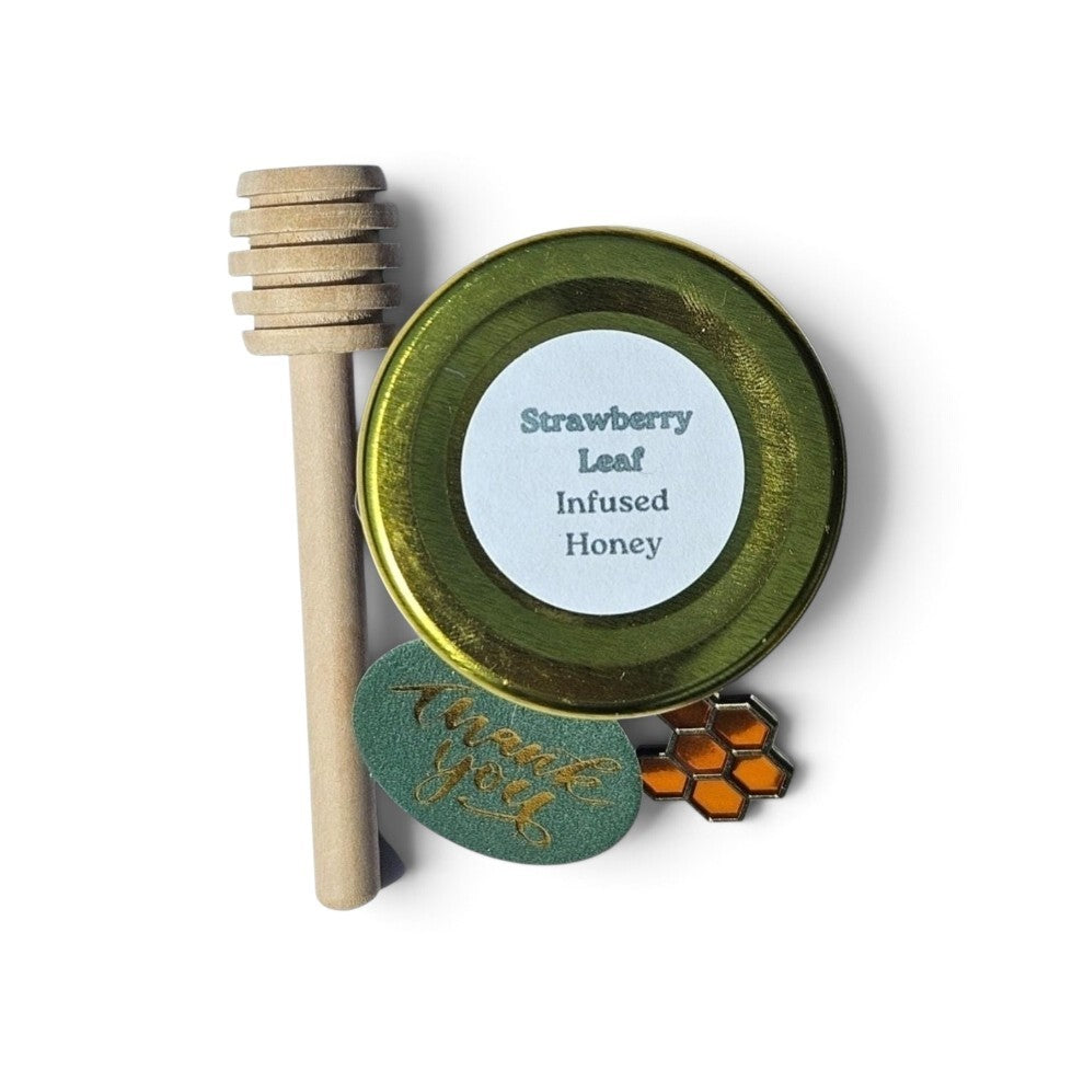 A jar of Strawberry Leaf Infused Honey with a wooden honey dipper, a green enamel pin, and a matching tag on a white background.