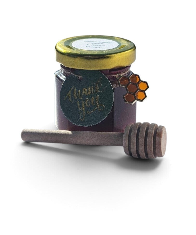 A small glass jar of dark purple honey with a gold lid and a round green 'thank you' label, shown with a wooden honey dipper on a white background.