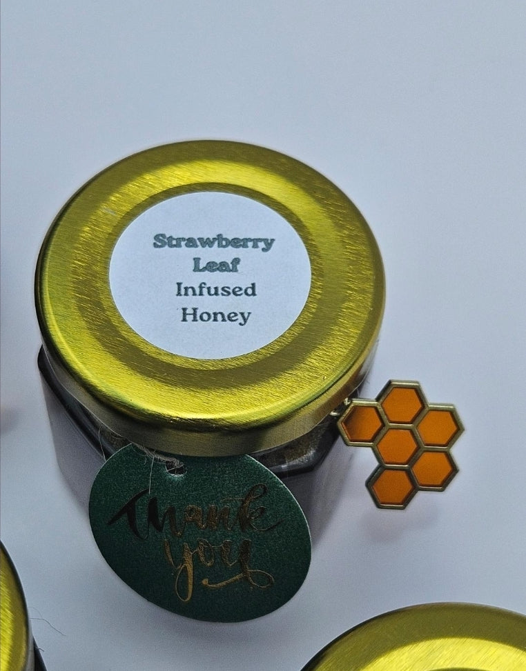 A small glass jar of strawberry leaf infused honey with a shiny gold lid and a white label. A green round tag reading 'thank you' hangs from the jar, and an orange honeycomb-shaped charm is attached to the side.