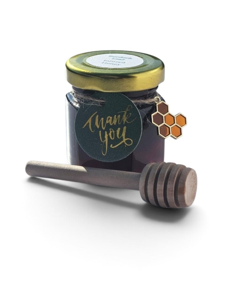 A small glass jar of dark honey with a gold metal lid and a round dark green 'thank you' tag, shown with a wooden honey dipper on a white background.