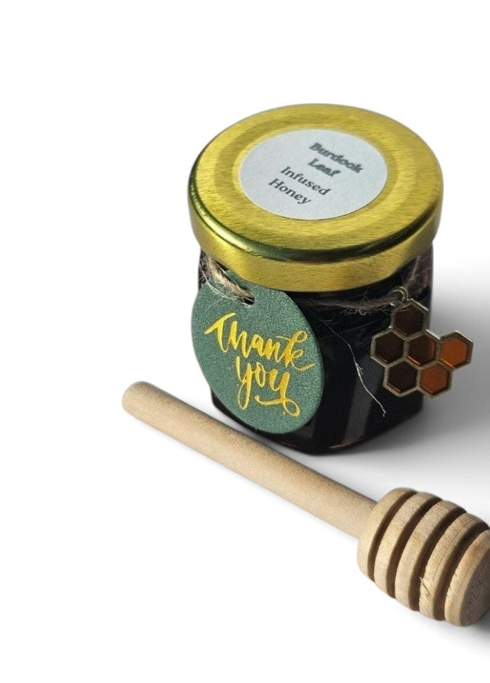 A jar of honey with a gold lid and a green 'thank you' tag, shown with a wooden honey dipper on a white background. The label reads 'Burdock Leaf Infused Honey'.